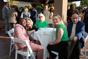 Five guests sit around a white-clothed round table at an outdoor daytime event, smiling for the camera.