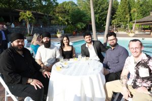 Six friends sit around a round table by a pool at an outdoor daytime event, with drinks and snacks on the table.