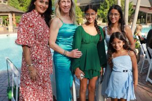 Five women and girls pose together by a pool at an outdoor event, wearing colorful summer dresses under palm trees.