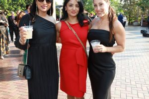 Three women in formal dresses posing together at an outdoor event, each holding a drink.
