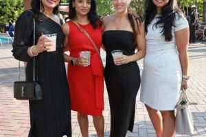 Four women in elegant dresses posing with drinks at an outdoor event, smiling for the camera.