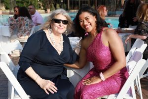 Two women sit at a table by a pool at an outdoor event, smiling for the camera; one wears a navy dress and sunglasses, the other a pink sequined dress hairstyle dark hair.