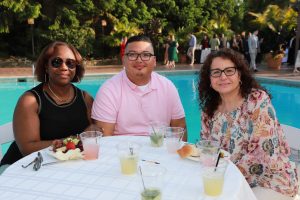 Three friends sit at a table by a pool, enjoying drinks and dessert at an outdoor gathering.