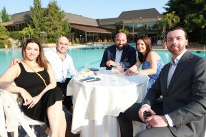 Five adults sit around a round table by a pool at an outdoor venue, smiling at the camera during a sunny event with drinks and plates nearby,