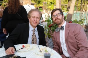 Two men in suits sit at an outdoor event, smiling at a round table with plates of dessert and a drink nearby.