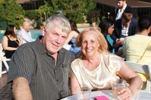 Smiling older couple seated at a table by a pool at an outdoor party, drinks in plastic cups nearby.