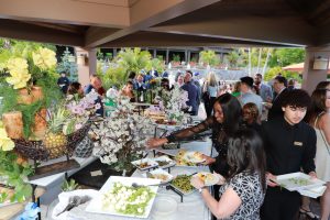Guests line up at a buffet table with assorted dishes and floral centerpieces at an outdoor event under a covered patio.