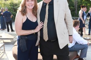 Couple posing together at an outdoor social event; woman in navy outfit with a wrap, man in beige blazer and patterned tie.