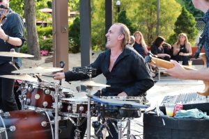 Drummer in a black shirt laughing while playing a red drum kit outdoors, with an audience seated behind him.