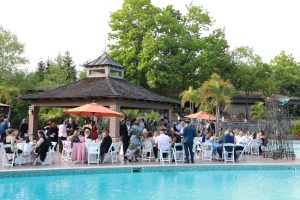 Guests mingle around round tables with white chairs and orange umbrellas by a pool, under a pavilion at an outdoor event.