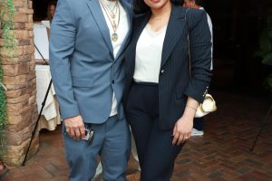 Man in a light blue suit and white sneakers poses with a woman in a navy pinstripe suit at an outdoor event, both looking at the camera and smiling.
