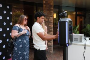 Two people stand at an outdoor event as a man uses a futuristic photo booth with a camera on top and glowing blue panels.