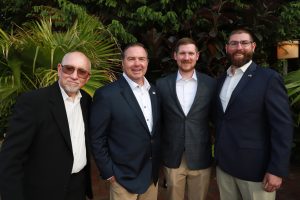 Four men in business attire posing together outdoors among greenery and plants, smiling at the camera.