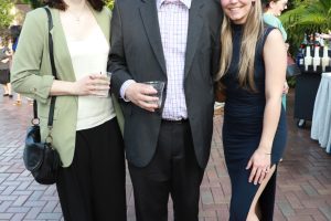 Three friends posing for a photo at an outdoor party; man in the center wears a suit, flanked by two women holding drinks and smiling.