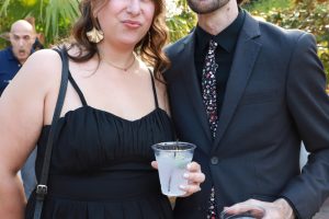 Couple standing outdoors in formal attire, each holding a drink and posing for the camera in a garden setting