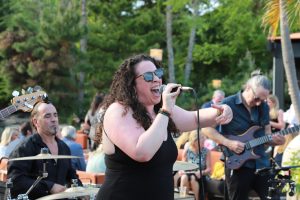 Female vocalist in black with sunglasses sings into a microphone on an outdoor stage, guitarist to her right and drummer to the left. Audience sits in the background.