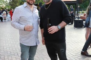 Two men pose together at an outdoor event, each holding a drink and smiling at the camera amid a courtyard party setting.