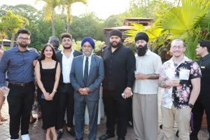 Eight people pose together at a sunny outdoor event, wearing smart casual outfits with drinks in hand, palm trees in the background.