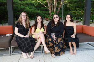 Four women sit on an outdoor sectional sofa, smiling, in a garden setting wearing dresses and sandals.