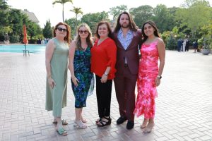 Group of five adults posing together at an outdoor poolside event, dressed in colorful outfits and smiling at the camera.
