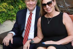 Smiling couple posing on a bench outdoors; man in a navy suit with a red striped tie and woman in a black dress with sunglasses.