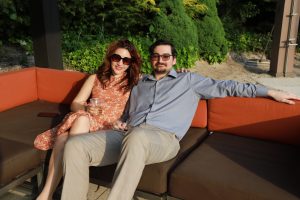 Couple relaxing on an outdoor brown and orange sectional couch, the woman in a peach floral dress and sunglasses sipping a drink while the man in a blue shirt leans back with his arm around her.