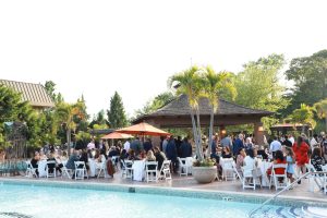 People gathered at a poolside garden party with white tables and chairs under orange umbrellas, a pavilion in the background.
