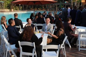 Group of formally dressed guests seated at round tables by a pool at an outdoor reception