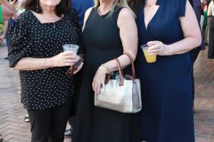 Three women pose together at an outdoor party; two hold drinks, one carries a metallic handbag on a brick patio.
