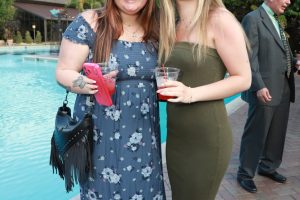 Two women pose by a pool at an outdoor gathering, each holding a drink; one in a blue floral dress with sunglasses, the other in an olive-green dress with sandals.