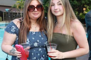 Two women standing by a pool during a social event, each holding a plastic cup with red drink; one wears a blue floral dress and sunglasses, the other a strapless olive dress.