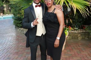 Couple in formal wear posing outdoors in a garden, with large palm fronds behind them.