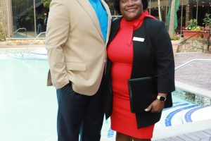 Man in a beige blazer and blue shirt stands beside a woman in a red dress and black blazer by a pool at an outdoor venue, smiling. It looks like a formal event setup.
