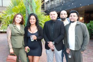 Group of five friends posing together at an outdoor event, smiling with green plants in the background.