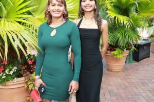 Two women pose outdoors among large tropical plants; one in a green dress, the other in a black dress.