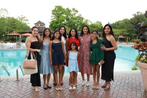Group of ten women and girls in dresses posing by a poolside, smiling for a photo at an outdoor gathering.