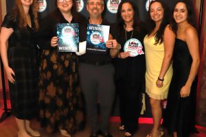Seven adults pose with winner plaques at a Long Island Choice Awards backdrop, smiling in formal attire.