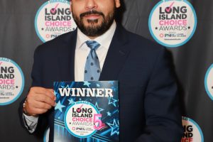 Man in a navy suit holding a 'WINNER' award certificate for the Long Island Choice Awards, posing in front of a themed backdrop.