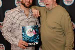 Two men pose for a photo at the Long Island Choice Awards backdrop; one holds a winner plaque.