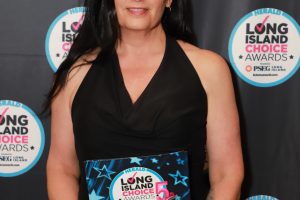 Woman in a black dress holding a Long Island Choice Awards winner certificate in front of an event backdrop with circular logos.