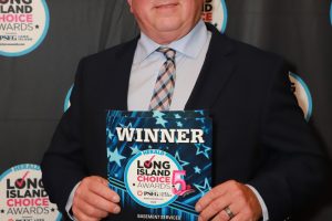 Man in a dark suit and tie holding a 'WINNER' plaque at the Long Island Choice Awards event, with backdrop of award logos.