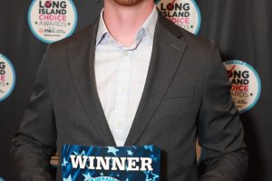 Man in a gray suit holding a 'WINNER' award plaque at the Long Island Choice Awards ceremony.