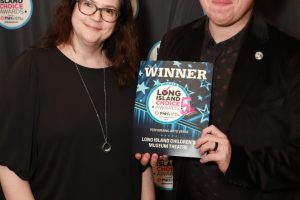 Two smiling adults pose in front of a backdrop covered with Long Island Choice Awards logos; the man holds a blue 'WINNER' plaque for the Long Island Children's Museum Theatre actions.