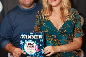 Man and woman smile at the camera while holding a blue 'WINNER' award plaque at the Long Island Choice Awards, standing in front of a backdrop with event logos.