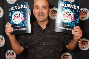Smiling man holding two 'WINNER' award certificates at a Long Island Choice Awards event, standing in front of a backdrop covered with circular logos.