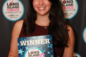 Smiling woman holding a blue 'WINNER' certificate at a Long Island Choice Awards backdrop.