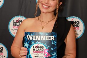 Smiling woman in a black dress holds a blue 'WINNER' certificate at the Long Island Choice Awards backdrop.