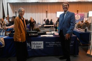 Two professionals at a conference booth for Meltzer Lippe LLP, with brochures and a navy table covered in branded materials behind them.