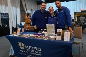 Three staff members in blue Metro Physical & Aquatic Therapy shirts stand behind a booth at a health fair, smiling at the camera with brochures on the table in front of them.