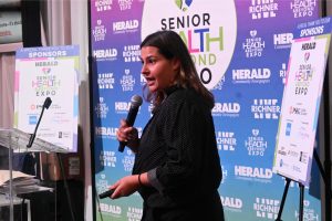 Woman speaking into a handheld microphone at a podium, with a blue expo backdrop and sponsor boards behind her.